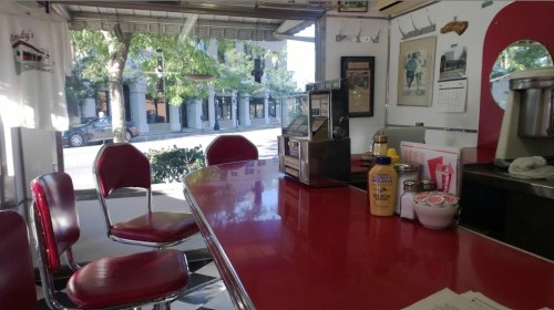 A retro diner interior with red stools and a shiny counter, overlooking a sunny street through large windows.