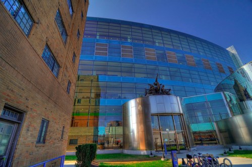 Modern glass building with a curved facade, featuring metallic accents and a sculpture at the entrance, surrounded by greenery.