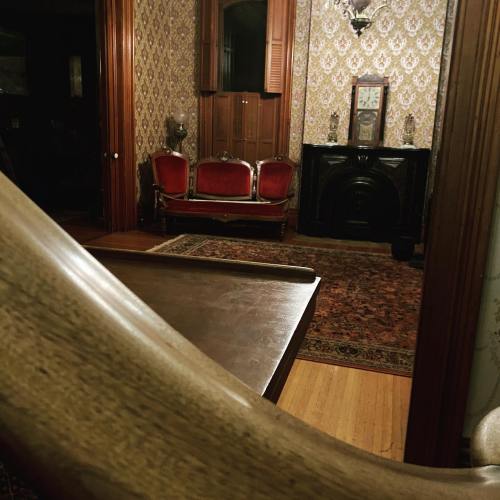 A vintage room with patterned wallpaper, a dark fireplace, red chairs, and wooden flooring, viewed from an angled perspective.