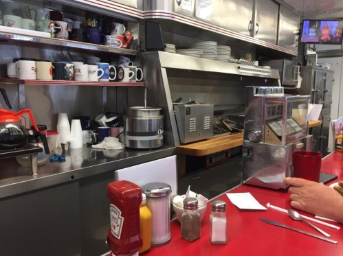 A diner counter with coffee mugs, condiments, and a jukebox, featuring a person’s hand resting on the counter.