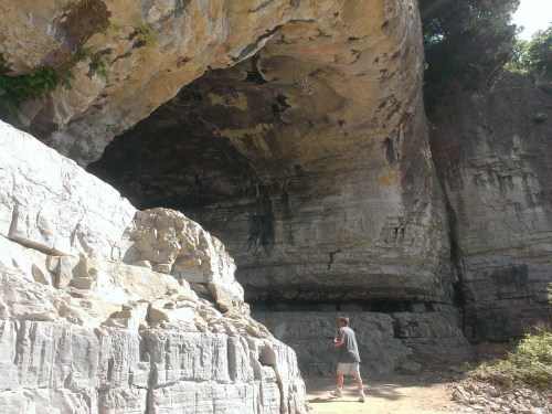 A person walks near a large rock formation with steep, textured cliffs and greenery above.