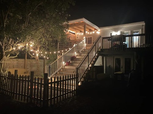 A well-lit backyard at night featuring a deck with stairs, string lights, and a fenced area.