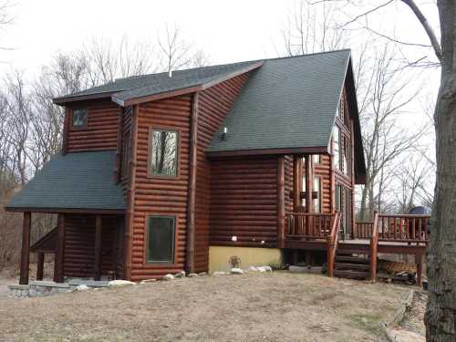 A large log cabin with a steep roof, surrounded by bare trees and a grassy area, featuring a wooden deck.