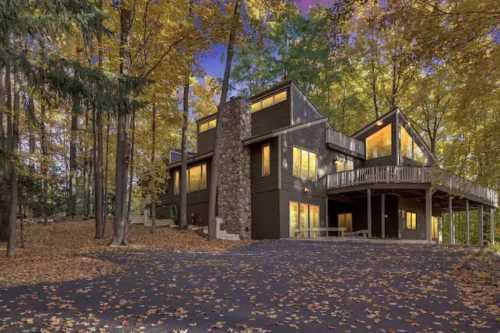 Modern house surrounded by autumn trees, featuring large windows and a stone chimney, with fallen leaves on the ground.