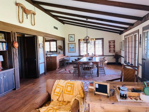 A cozy, rustic dining area with wooden beams, a large table, and warm decor, featuring a sofa and decorative items.