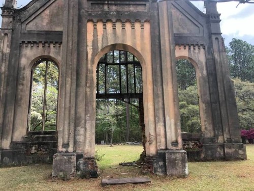 Ruins of a stone structure with arched windows, surrounded by greenery and trees under a cloudy sky.