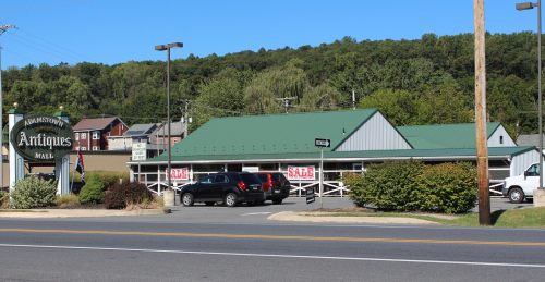 A view of an antique mall with a green roof, surrounded by trees and parked cars, along a road.