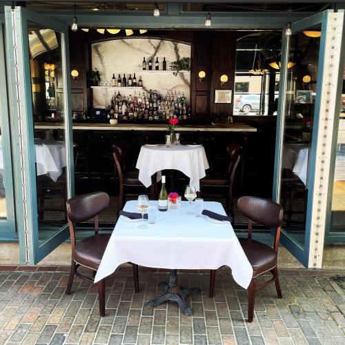 A cozy restaurant with two empty tables set for dining, featuring a bar in the background and outdoor seating.