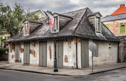 Historic building with a weathered exterior, featuring shutters, an American flag, and a lamppost on a quiet street.