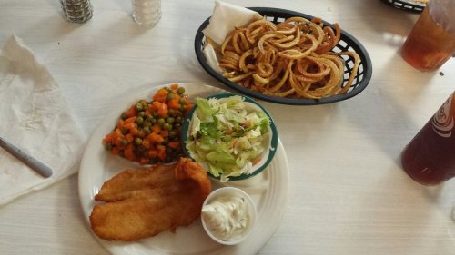 A plate with fried fish, mixed vegetables, salad, and tartar sauce, alongside a basket of curly fries.