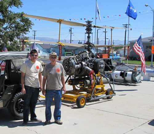 Two men stand beside a helicopter engine on a yellow cart, with flags and a military vehicle in the background.