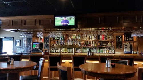 A cozy bar with wooden tables, a well-stocked shelf of liquor, and a TV showing a program in a dimly lit setting.