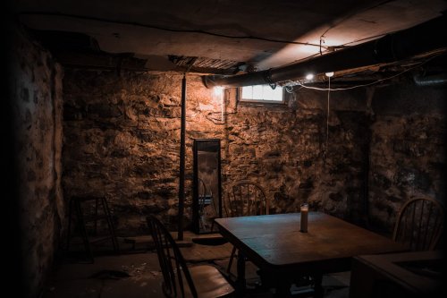 Dimly lit basement with stone walls, a wooden table, chairs, and a mirror reflecting light from a small window.