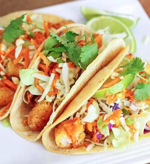 Three tacos filled with crispy fish, shredded cabbage, carrots, and garnished with lime and cilantro on a white plate.