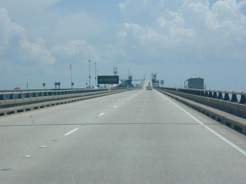 A wide highway stretches ahead, leading to a bridge with a sign and structures under a cloudy sky.