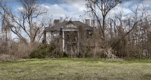 An abandoned, overgrown mansion surrounded by trees and brush under a cloudy sky.