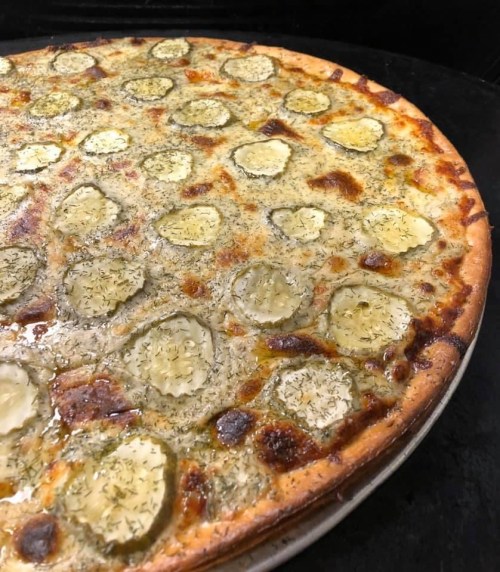 A pizza topped with pickles and herbs, featuring a golden crust and a cheesy, bubbly surface.