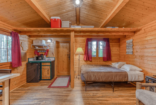 Cozy wooden cabin interior with a bed, kitchenette, and warm lighting, surrounded by windows and rustic decor.