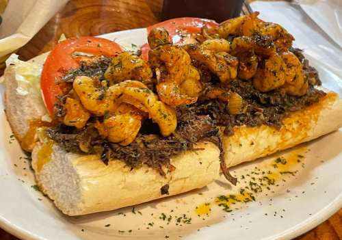 A loaded sandwich with shrimp, tomatoes, and greens on a toasted hoagie roll, garnished with herbs.