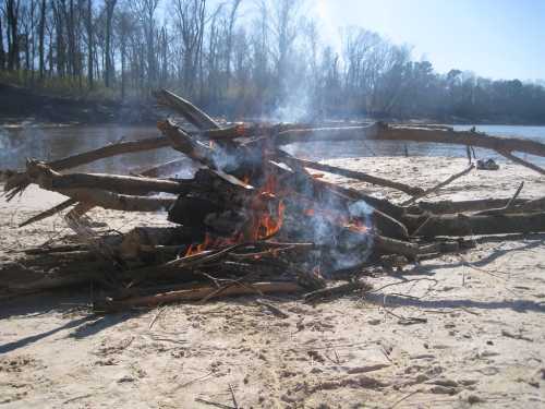 A campfire made of logs burns on a sandy riverbank, with smoke rising against a backdrop of bare trees.