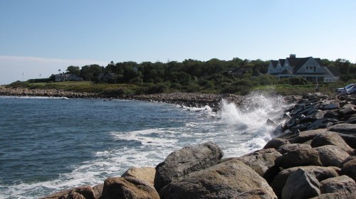 Waves crash against rocky shore with a coastal house in the background under a clear blue sky.