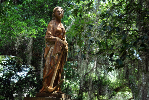 A wooden statue of a woman holding an anchor, surrounded by lush greenery and hanging moss.