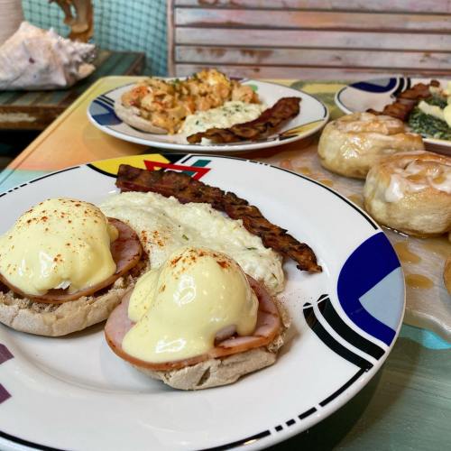 A plate of eggs Benedict with ham and hollandaise sauce, alongside bacon and other breakfast dishes in a colorful setting.
