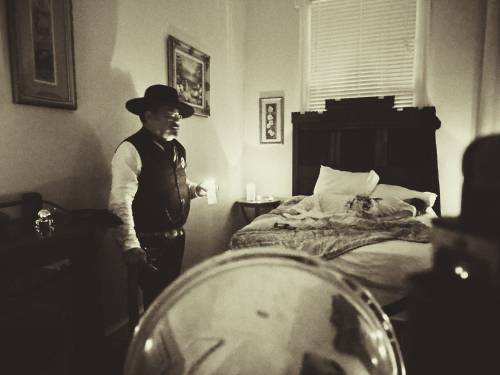 A man in a hat stands in a dimly lit bedroom, holding a candle near a bed with a quilt.