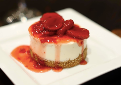 A creamy cheesecake topped with strawberries and a glossy red sauce, served on a white plate.