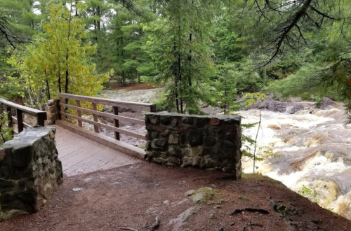 A wooden bridge over a rushing river, surrounded by lush green trees and rocky terrain.