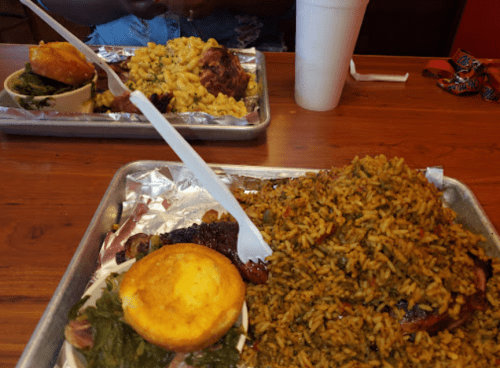 Two trays of food featuring rice, cornbread, macaroni and cheese, greens, and a meat dish, served in a casual dining setting.