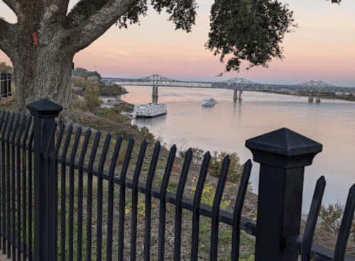 A serene river view at sunset, featuring a bridge and a boat, framed by a tree and a black fence.