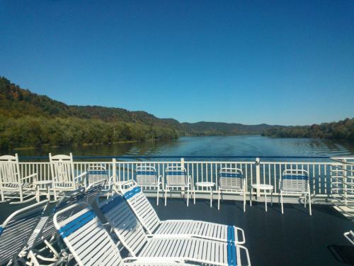 View from a boat deck with white chairs overlooking a calm river and green hills under a clear blue sky.