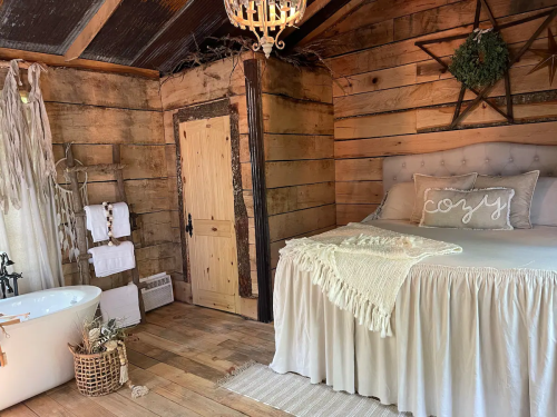 Cozy rustic interior featuring a bed, bathtub, wooden walls, and decorative elements like a chandelier and wreath.