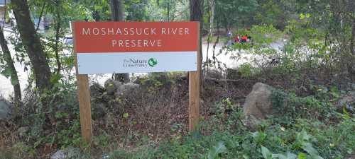 Sign for Moshasuck River Preserve, surrounded by trees and greenery, with people visible in the background.