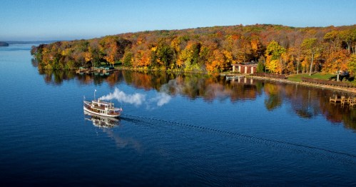 A steamboat glides through a calm river, surrounded by vibrant autumn foliage and a scenic shoreline.