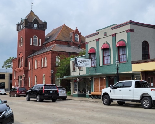 Historic buildings line a street, featuring a clock tower and a restaurant with red awnings. Cars are parked along the road.