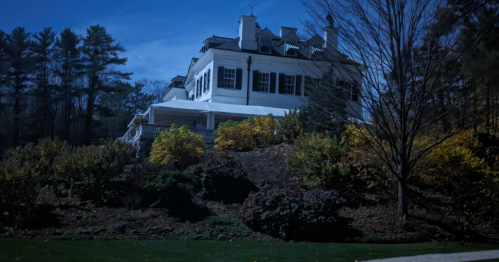A large, white house sits atop a hill, surrounded by trees and autumn foliage under a clear blue sky.