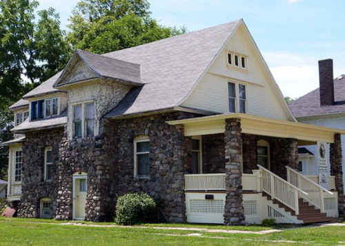 A large, two-story house with a stone exterior, a porch, and a gabled roof, surrounded by greenery.