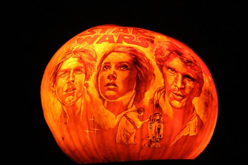 A carved pumpkin featuring iconic characters from Star Wars, illuminated in orange light against a dark background.