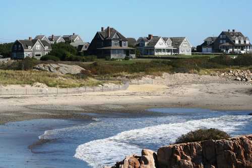 Coastal view of beach with gentle waves and a row of large houses on a grassy hill in the background.