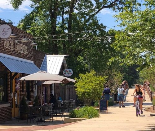 A sunny street scene with a coffee shop, outdoor seating, and people walking and biking along a path.