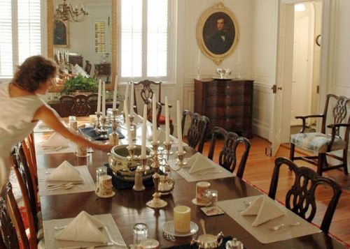 A woman sets a formal dining table with candles and silverware in an elegant room with vintage decor.