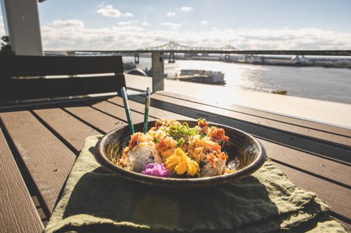A plate of sushi with colorful garnishes sits on a table overlooking a river and bridge under a clear sky.