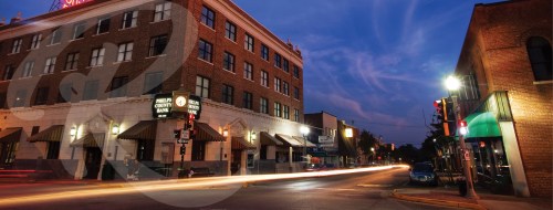 A street view at dusk featuring a brick building, traffic lights, and light trails from passing cars.