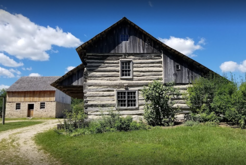 A rustic log cabin and a stone building surrounded by greenery under a blue sky with fluffy clouds.