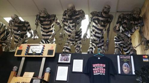 Five skeletons in striped prison uniforms hang from the ceiling, with a display of a T-shirt and framed documents below.