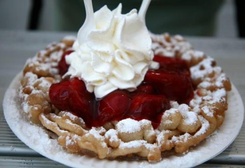 A plate of funnel cake topped with whipped cream and strawberries, dusted with powdered sugar.