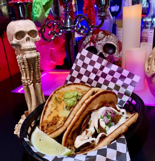 Three tacos in a basket with lime, surrounded by decorative skulls and colorful lighting.