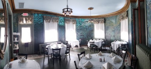 A cozy dining room with round tables, white tablecloths, and tropical-themed wallpaper, featuring large windows and elegant decor.